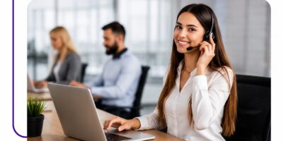 Customer service rep wearing a headset and typing on a laptop in an open office. Two colleagues in the background are blurred.