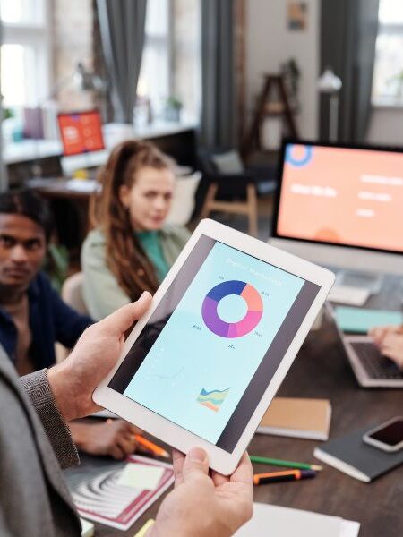 Person holding a tablet showing a colorful pie chart during a meeting with coworkers in a bright office.